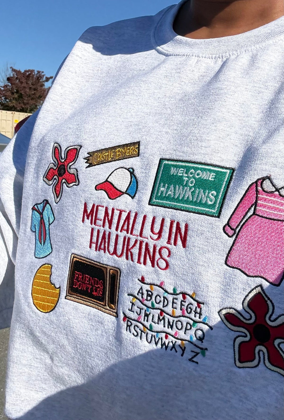 A close-up of a person wearing a light gray crewneck sweatshirt with a colorful embroidery on the front featuring the text 'MENTALLY IN HAWKINS' and various tv show themed designs