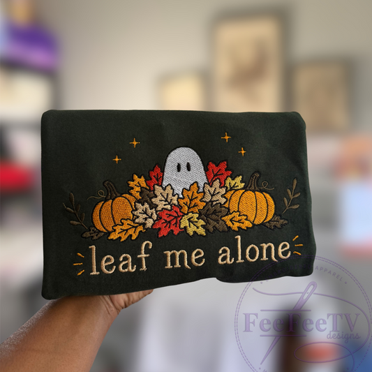 A hand holds a forest green tshirt featuring a Halloween-themed embroidered design with a white ghost, pumpkins, autumn leaves, and the phrase 'leaf me alone' in gold thread. The background is a blurred indoor setting.