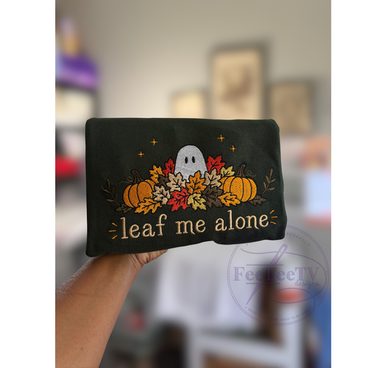 A hand holds a forest green sweatshirt featuring a Halloween-themed embroidered design with a white ghost, pumpkins, autumn leaves, and the phrase 'leaf me alone' in gold thread. The background is a blurred indoor setting.