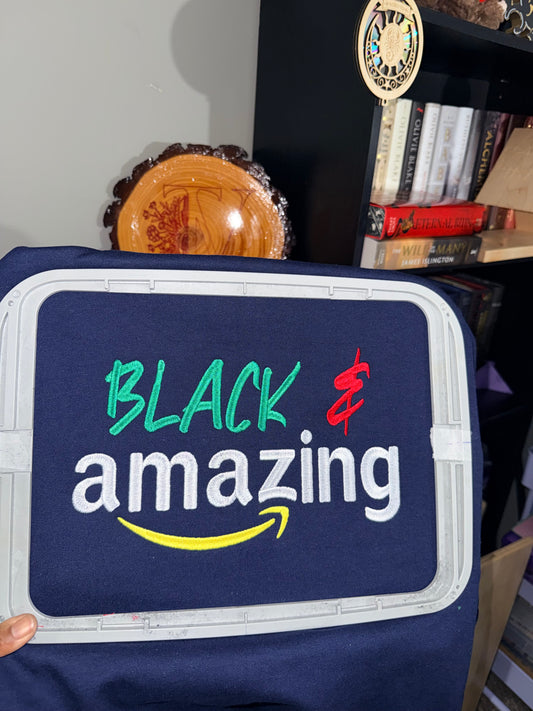 Navy blue sweatshirt secured in an embroidery hoop featuring a “Black & amazing” design stitched in green, red, white, and yellow thread, shown in a home studio setting with bookshelves in the background.