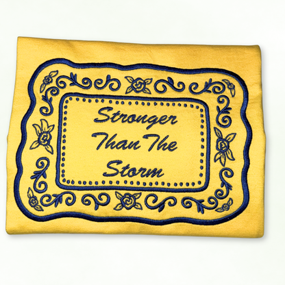 Yellow tshirt with blue embroidery and 'Stronger Than The Storm' text on a white background