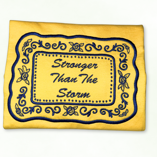 Yellow tshirt with blue embroidery and 'Stronger Than The Storm' text on a white background