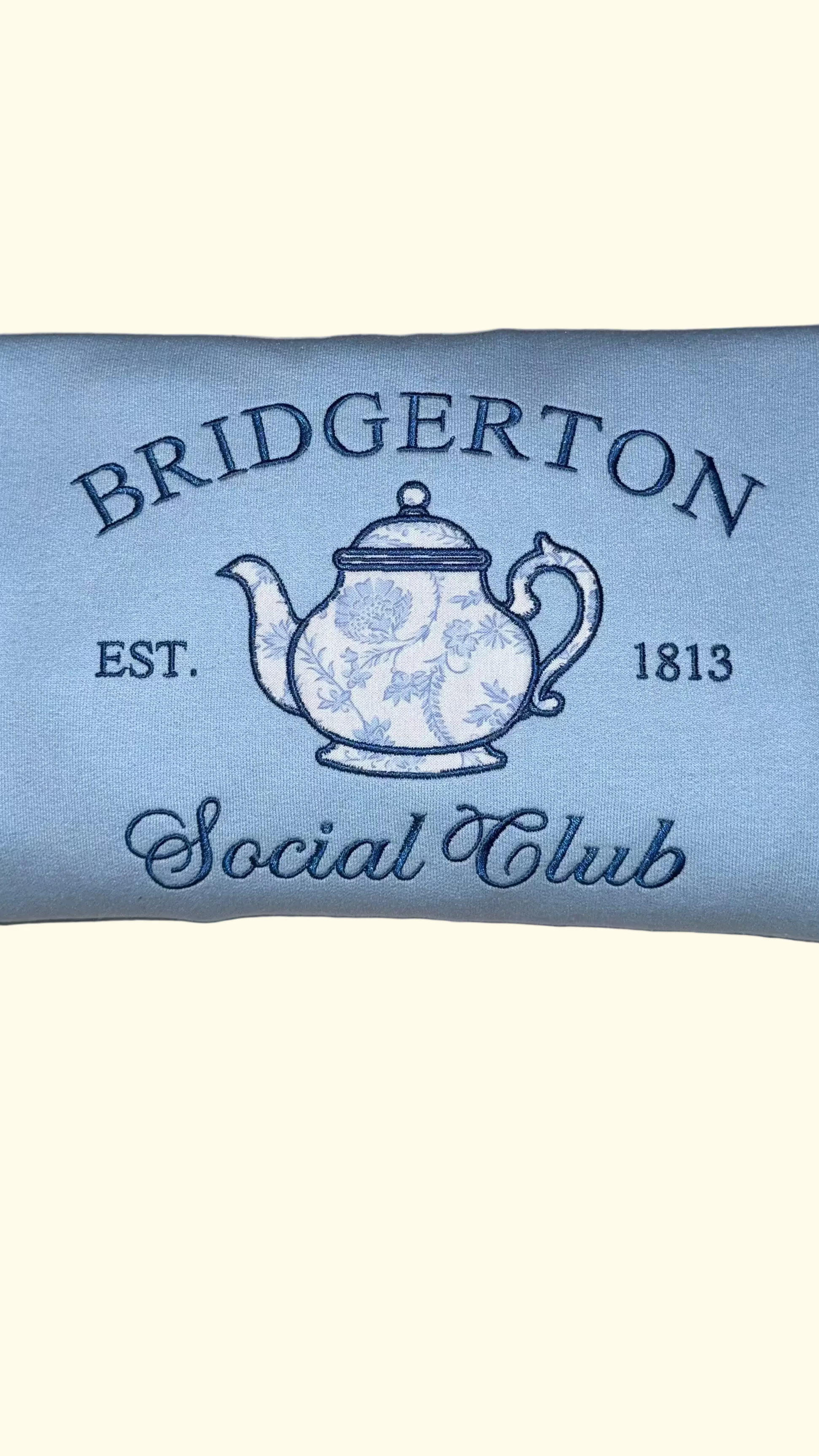 Close up of Bridgerton Social Club sweatshirt design featuring teapot illustration and vintage lettering on light blue crewneck