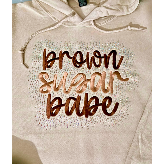 A close-up of a sand-colored hoodie featuring the phrase 'brown sugar babe' surrounded by  iridescent rhinestone bling