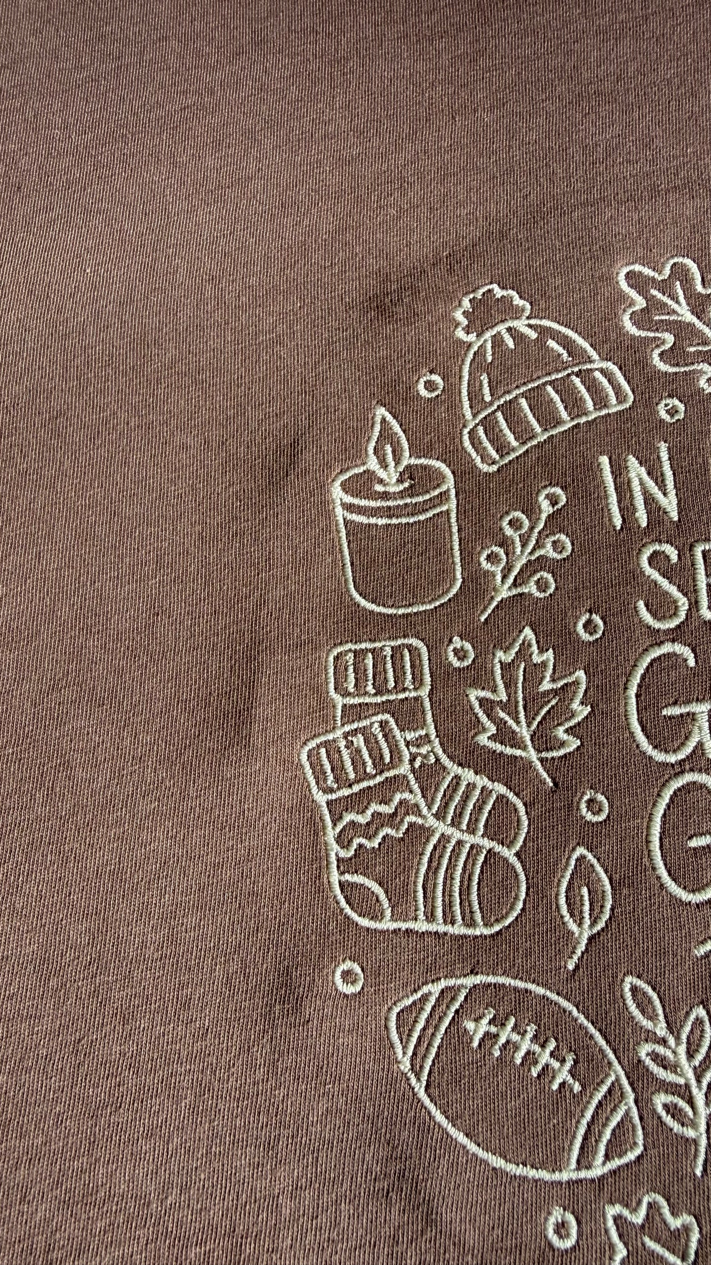 Close-up of embroidery on a brown t-shirt with cream outlines of a beanie, candle, socks, leaves, football.