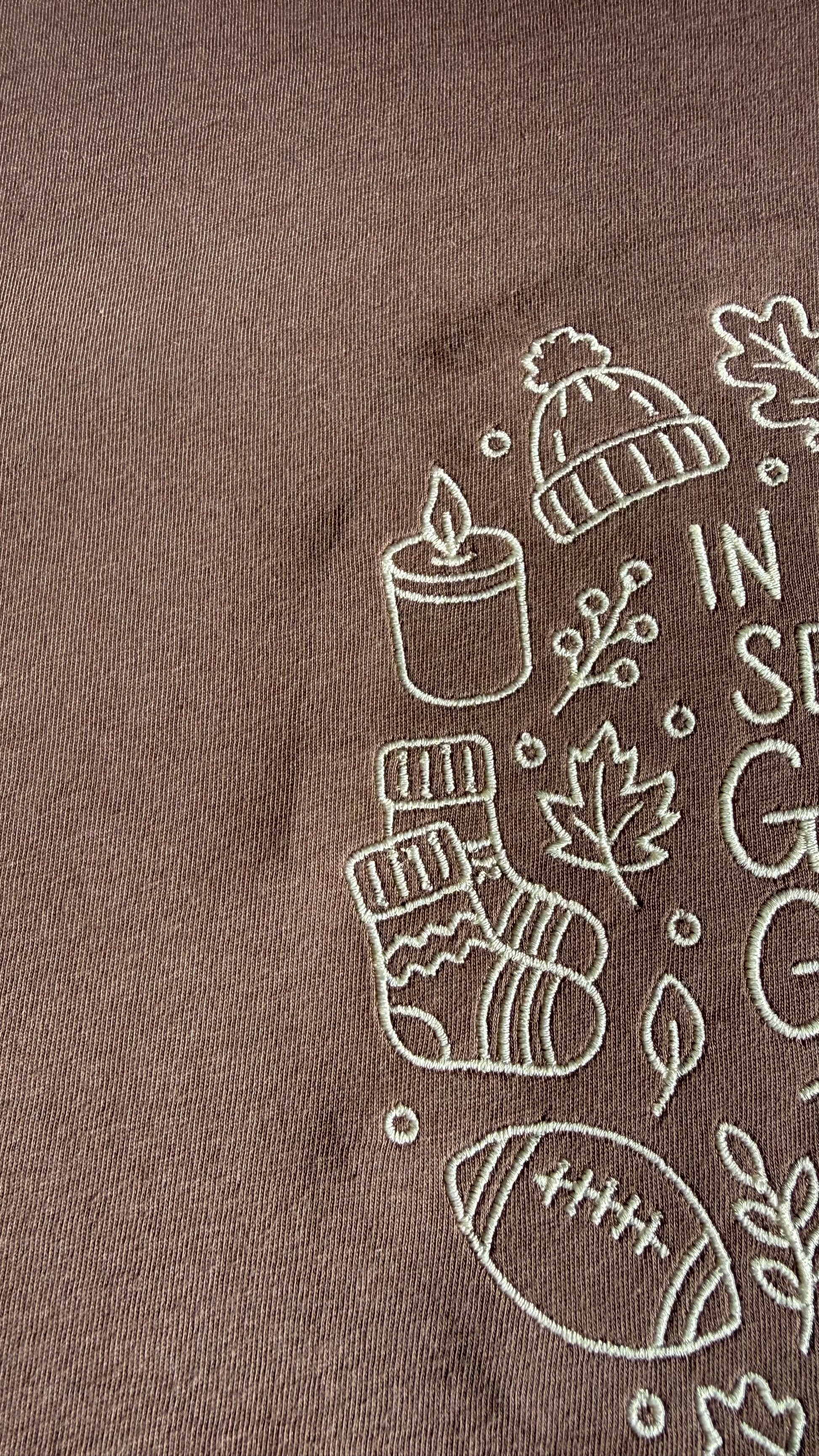 Close-up of embroidery on a brown t-shirt with cream outlines of a beanie, candle, socks, leaves, football.