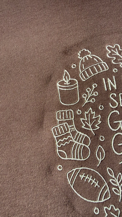 Close-up of embroidery on a brown t-shirt with cream outlines of a beanie, candle, socks, leaves, football.