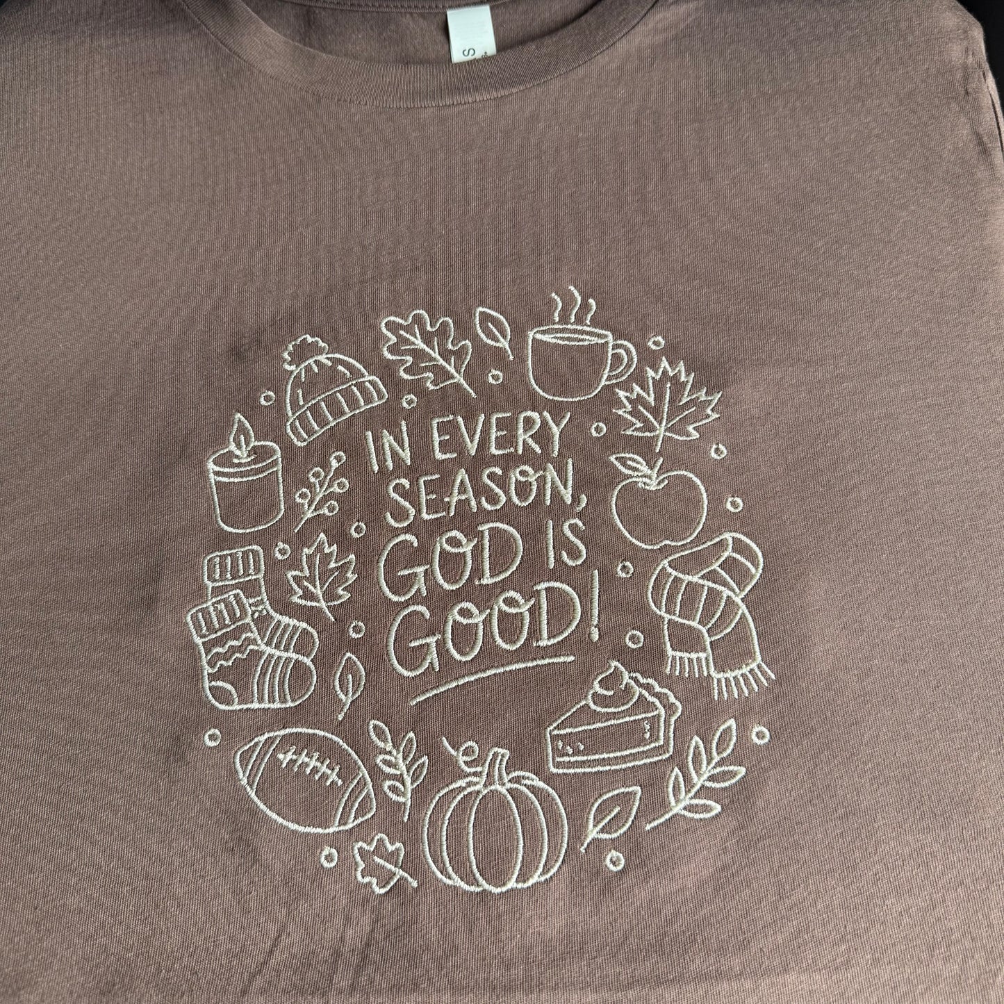A close-up of a brown crewneck t-shirt featuring a white embroidered graphic of autumn and winter elements surrounding the text 'IN EVERY SEASON, GOD IS GOOD!'.
