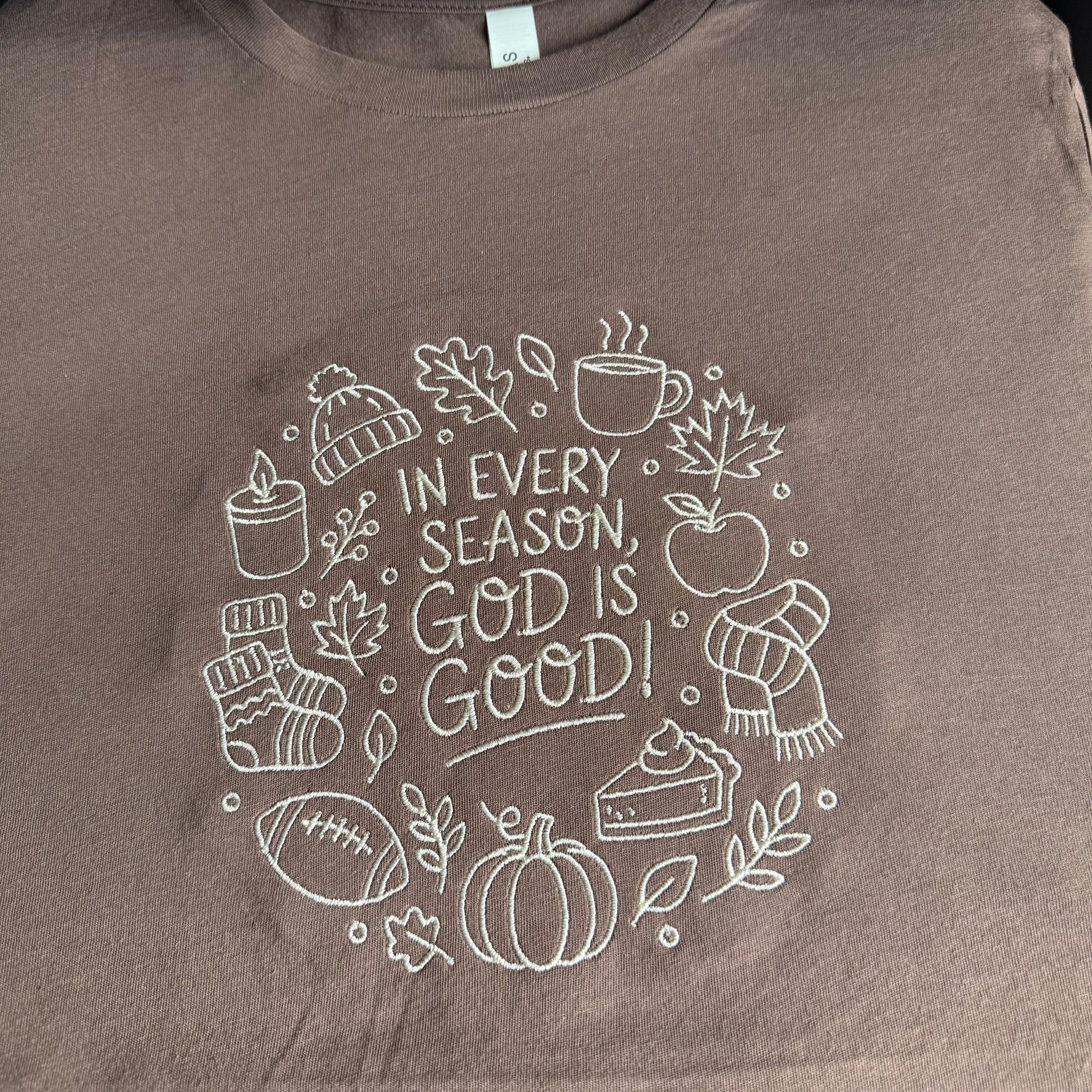 A close-up of a brown crewneck t-shirt featuring a white embroidered graphic of autumn and winter elements surrounding the text 'IN EVERY SEASON, GOD IS GOOD!'.
