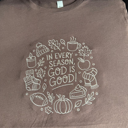 A close-up of a brown crewneck t-shirt featuring a white embroidered graphic of autumn and winter elements surrounding the text 'IN EVERY SEASON, GOD IS GOOD!'.