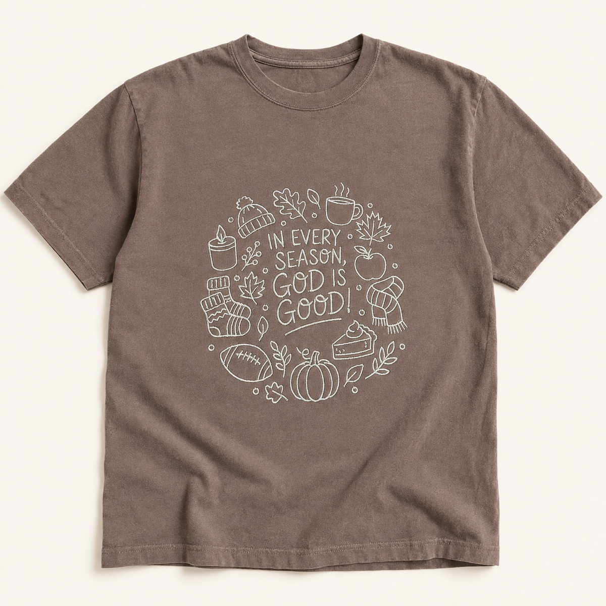 A brown short-sleeve crewneck t-shirt with a cream embroidery on the chest containing autumn-themed icons and the text 'IN EVERY SEASON, GOD IS GOOD'.