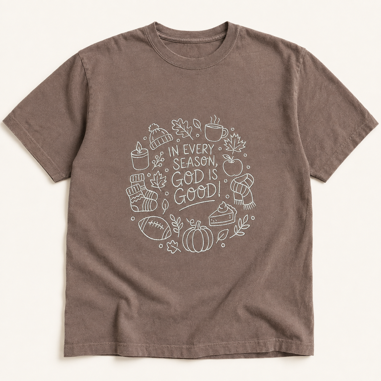 A brown short-sleeve crewneck t-shirt with a cream embroidery on the chest containing autumn-themed icons and the text 'IN EVERY SEASON, GOD IS GOOD'.