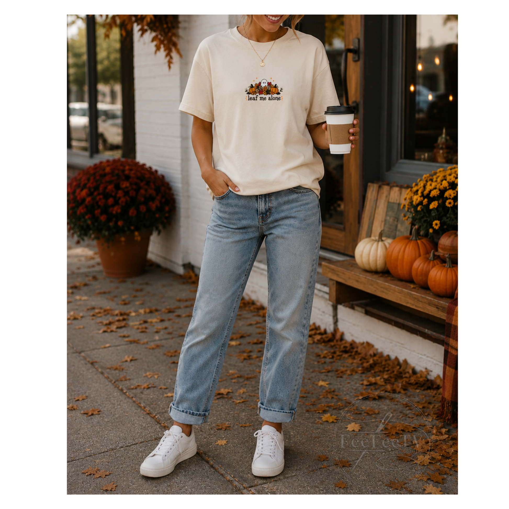A woman stands outdoors on a sidewalk wearing a cream embroidered T-shirt with a small autumn-themed print, light blue jeans, and white sneakers, while holding a coffee cup. Pumpkins, mums, and fallen leaves are visible around a storefront.