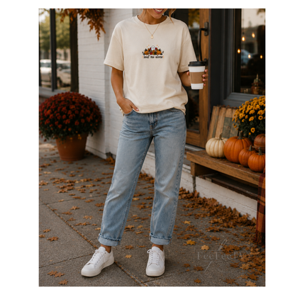 A woman stands outdoors on a sidewalk wearing a cream embroidered T-shirt with a small autumn-themed print, light blue jeans, and white sneakers, while holding a coffee cup. Pumpkins, mums, and fallen leaves are visible around a storefront.