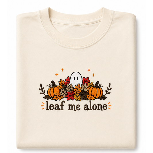 A folded cream-colored crewneck featuring a Halloween graphic with pumpkins, autumn leaves, a ghost, and the text 'leaf me alone' in brown, orange, and red.