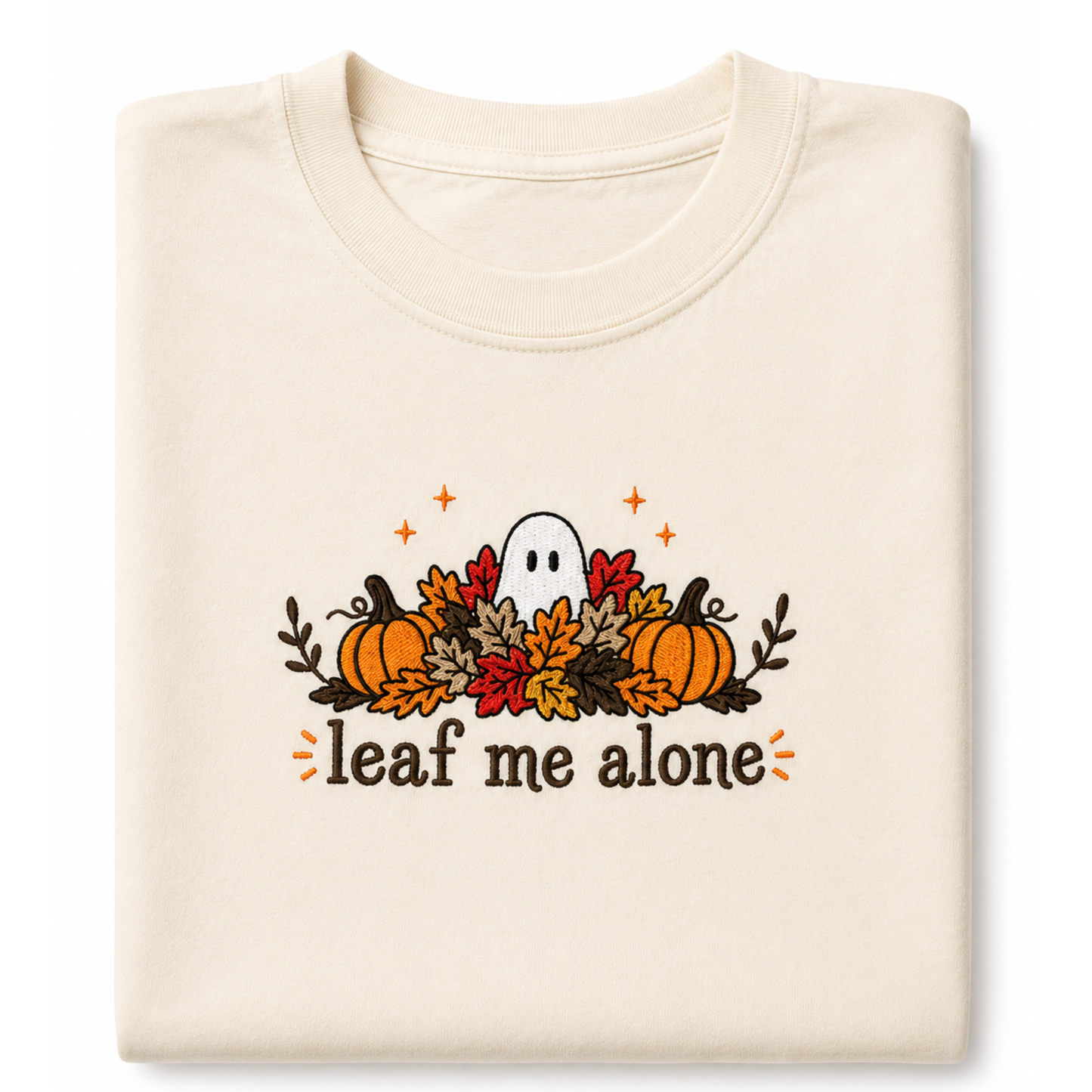 A folded cream-colored t-shirt featuring a Halloween graphic with pumpkins, autumn leaves, a ghost, and the text 'leaf me alone' in brown, orange, and red.