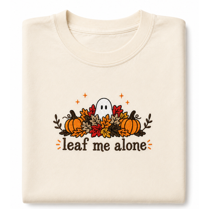 A folded cream-colored t-shirt featuring a Halloween graphic with pumpkins, autumn leaves, a ghost, and the text 'leaf me alone' in brown, orange, and red.