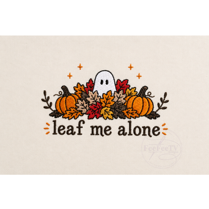 A embroidery design on a light beige fabric background, featuring a white ghost surrounded by orange, red, and yellow autumn leaves, two pumpkins, and the text 'leaf me alone'.