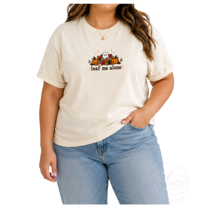 A woman wearing a cream-colored short-sleeve t-shirt with a fall-themed embroidery of pumpkins, a ghost, and the text 'leaf me alone', paired with light-wash blue jeans, against a plain white background.