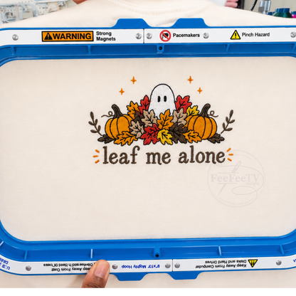 An embroidery hoop holds a white fabric with a completed autumn-themed embroidery design. The design shows orange pumpkins, red and yellow leaves, a small white ghost, and the words “leaf me alone” in brown.