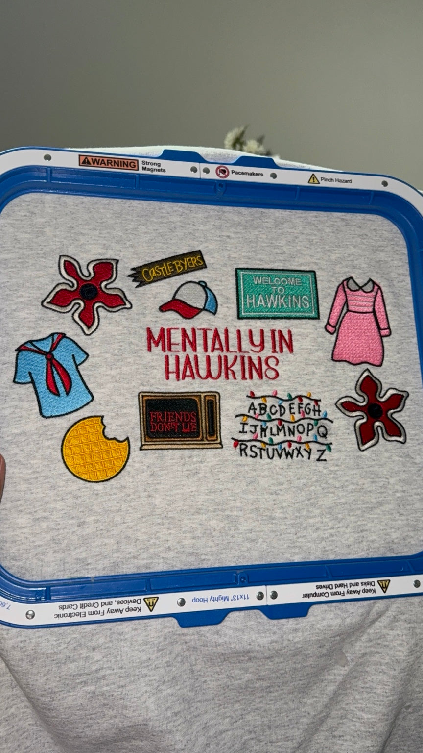 Gray crewneck sweatshirt featuring colorful embroidery and Mentally in Hawkins lettering, shown during embroidery.