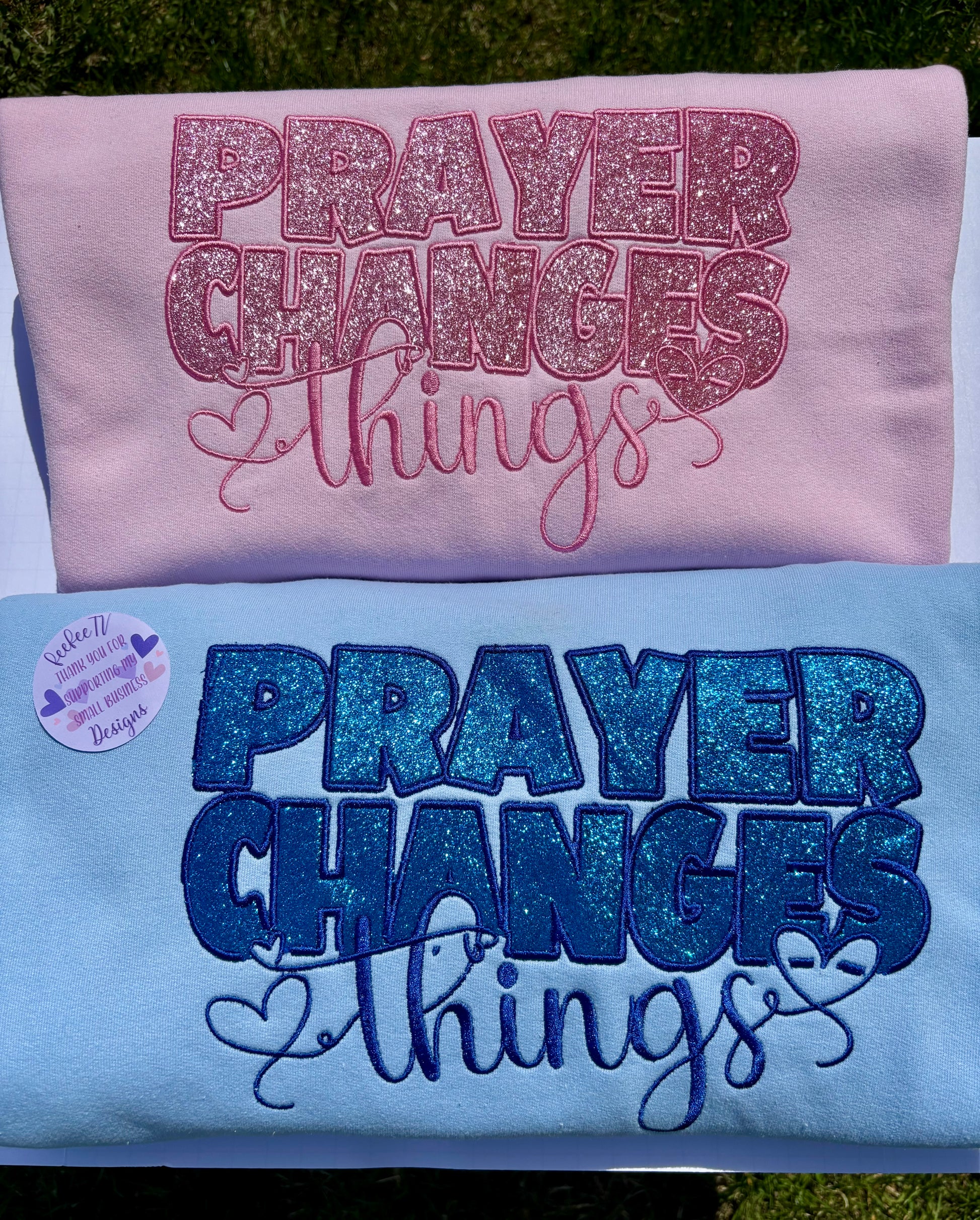 Two sweatshirts, one pink and one blue, each with the embroidered text 'PRAYER CHANGES THINGS' accompanied by heart shapes and other designs in glitter.