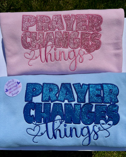 Two sweatshirts, one pink and one blue, each with the embroidered text 'PRAYER CHANGES THINGS' accompanied by heart shapes and other designs in glitter.