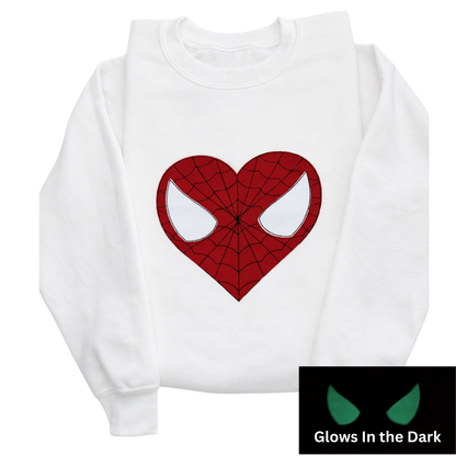 A white long-sleeve sweatshirt featuring a red spider web-heart and glow in the dark mask eyes. A small inset in the bottom right corner shows the graphic glowing green in the dark.